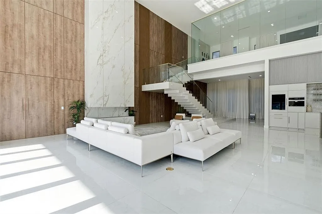 Living room with white sectional sofas, a full-height wood wall, and a floating staircase rising to a glass-walled loft.
