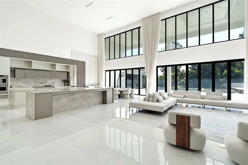 Kitchen with a long stone waterfall island facing a two-story wall of black-framed windows and a white sectional sofa.