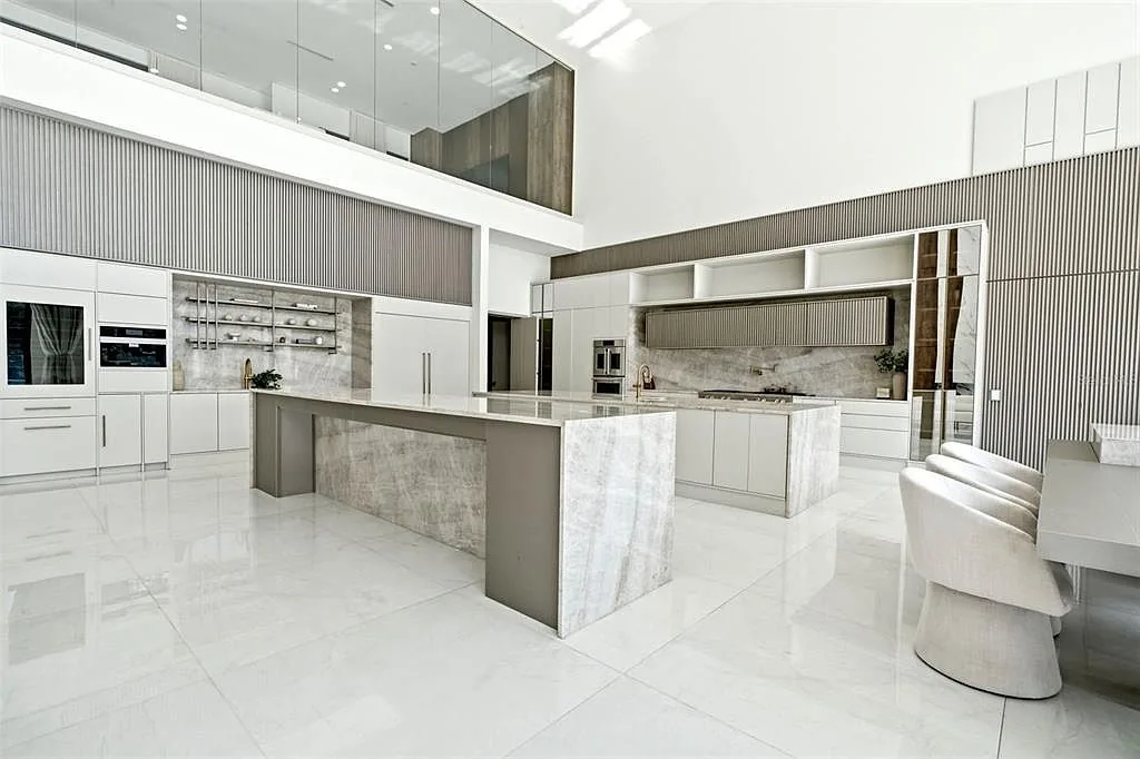 Kitchen with two stone waterfall islands, fluted wall panels, and upholstered dining chairs below a glass-walled loft.