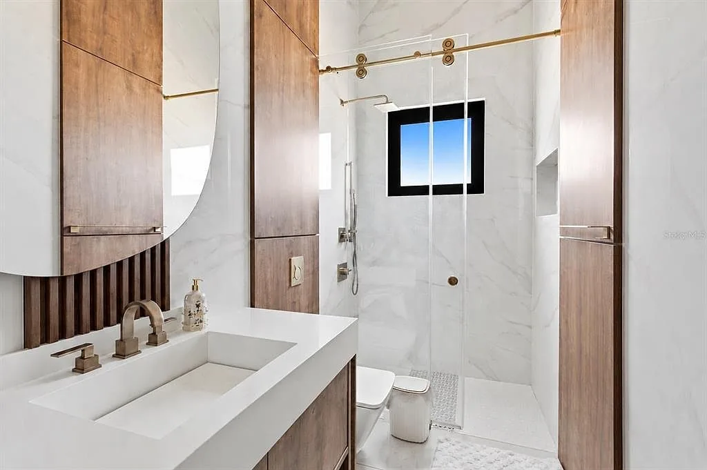 Bathroom with a walnut vanity, a curved mirror, and a marble-walled shower with brass fixtures.