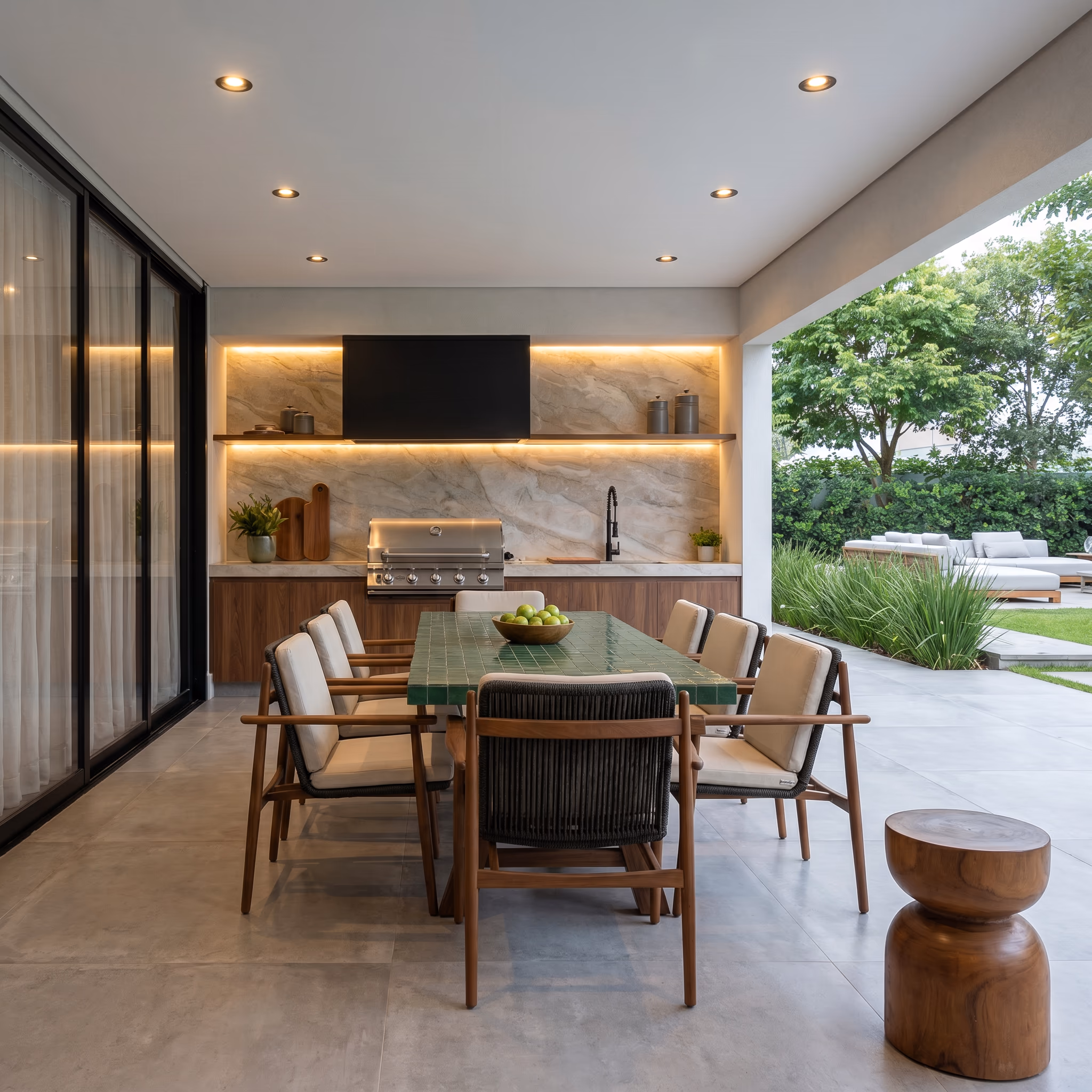 Covered summer kitchen with a built-in grill, a lit stone backsplash, a green-topped dining table with wood chairs, and open sliding doors to the garden.